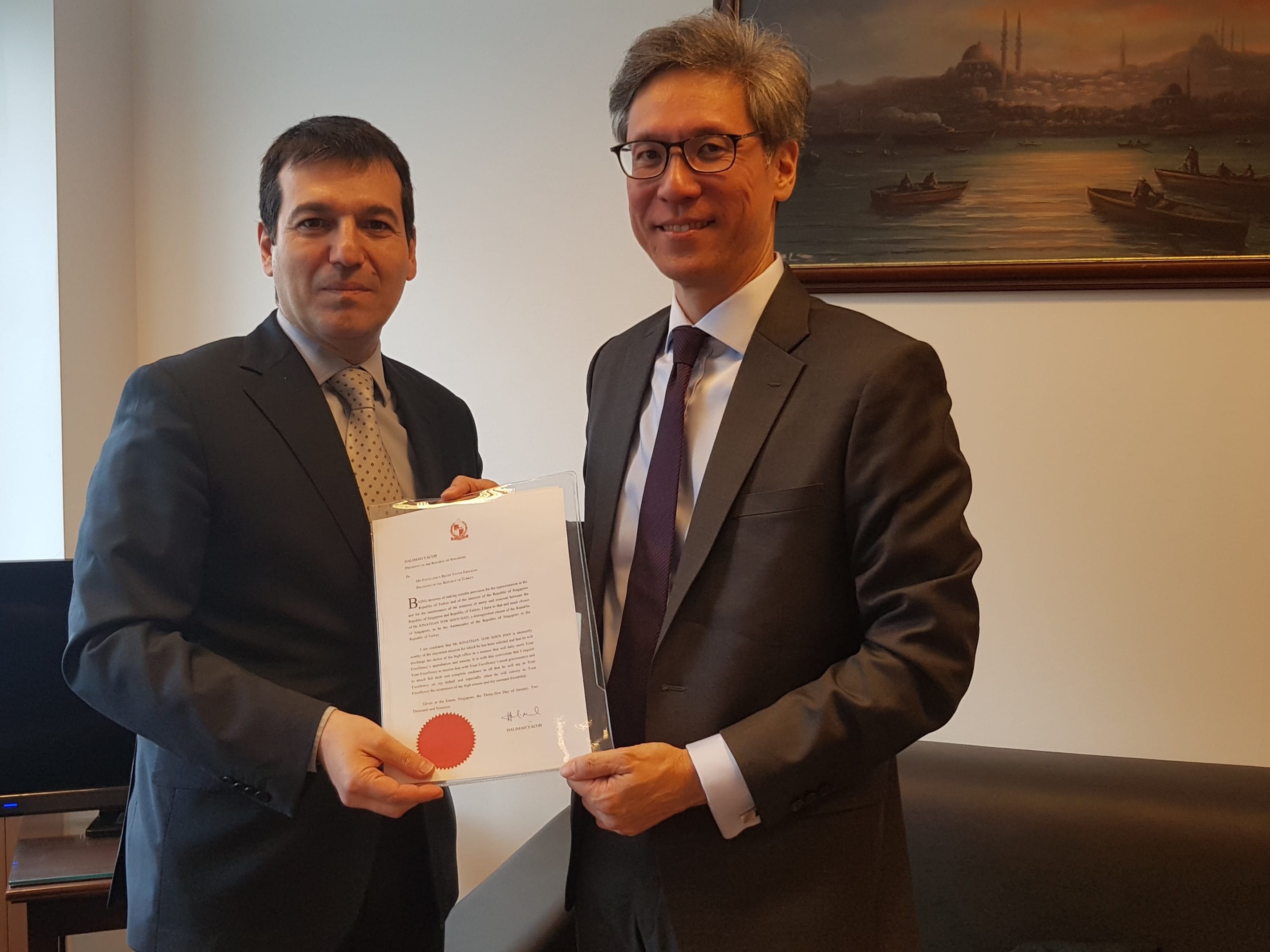 Two men in suits hold a document with a red seal. A painting is visible in the background.